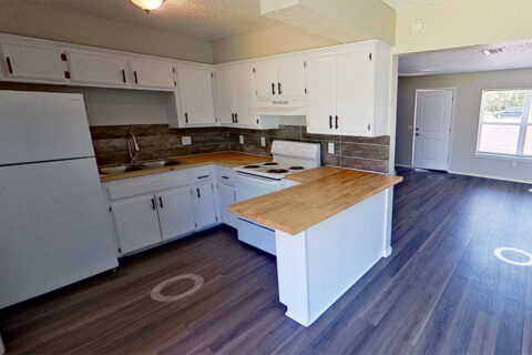 A kitchen with white cabinets, wood countertops, a white refrigerator, and an electric stove. The room has wood-look flooring and an open layout leading to a doorway and windows.